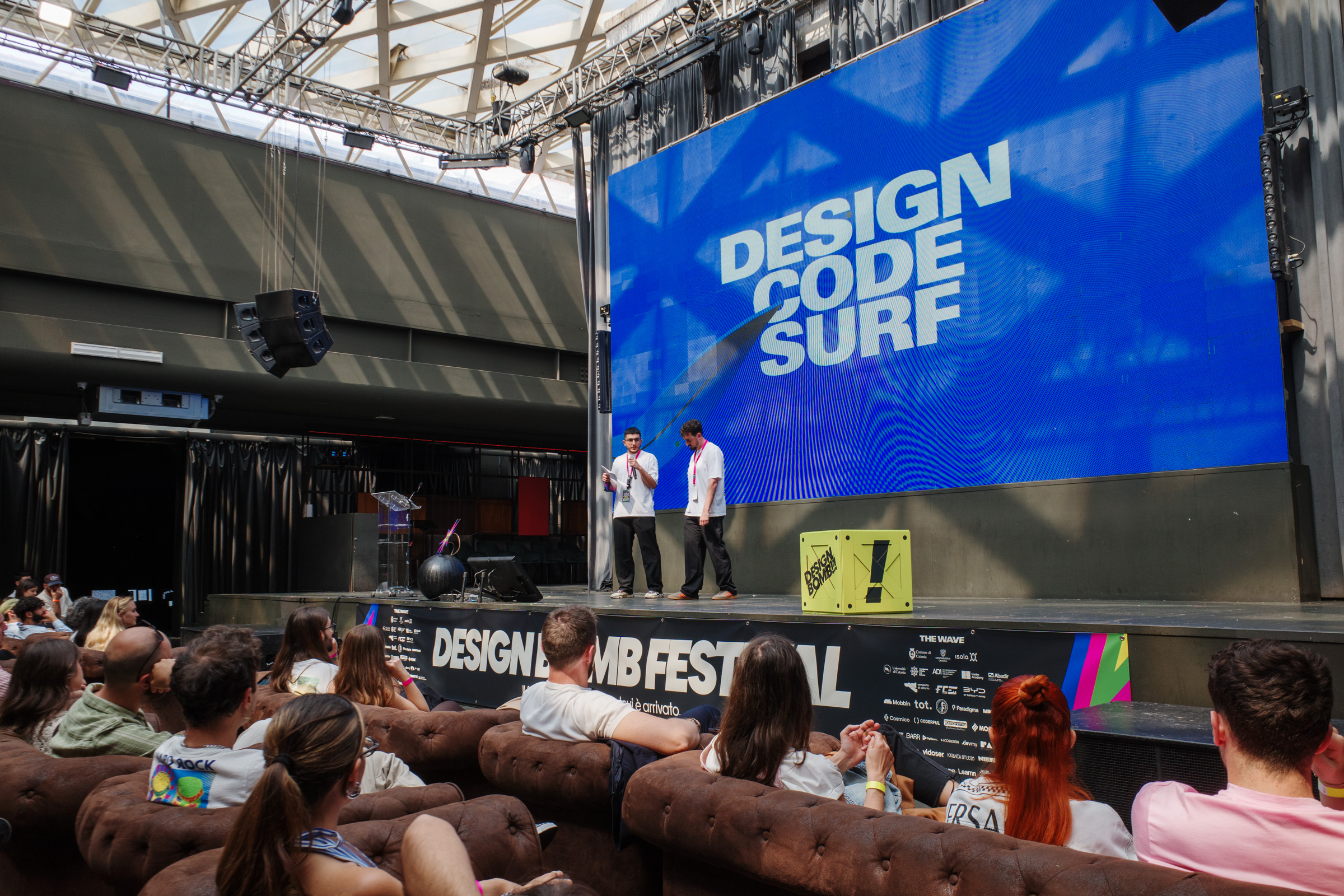 Il palco del Design Bomb Festival con due speaker e lo schermo con la scritta Design Code Surf