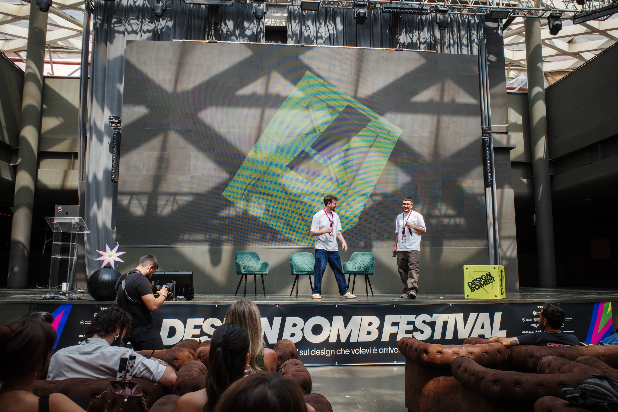 Due organizzatori sul palco del Design Bomb Festival con uno schermo grafico sullo sfondo